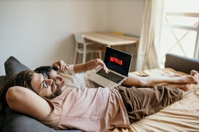 Man in white t-shirt lying on bed using macbook to stream.
