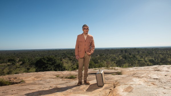 Eugene Levy steht auf einem Felsen mit Aussicht auf ene grne Landschaft, neben ihm ein Koffer